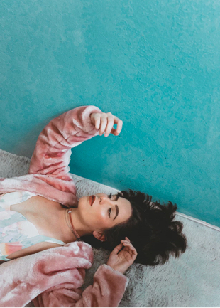 Harper laying down on her grey faux fur rug looking thoughtfully up at her fingernails. She is wearing her favourite pink faux fur coat over a sleek summer dress.